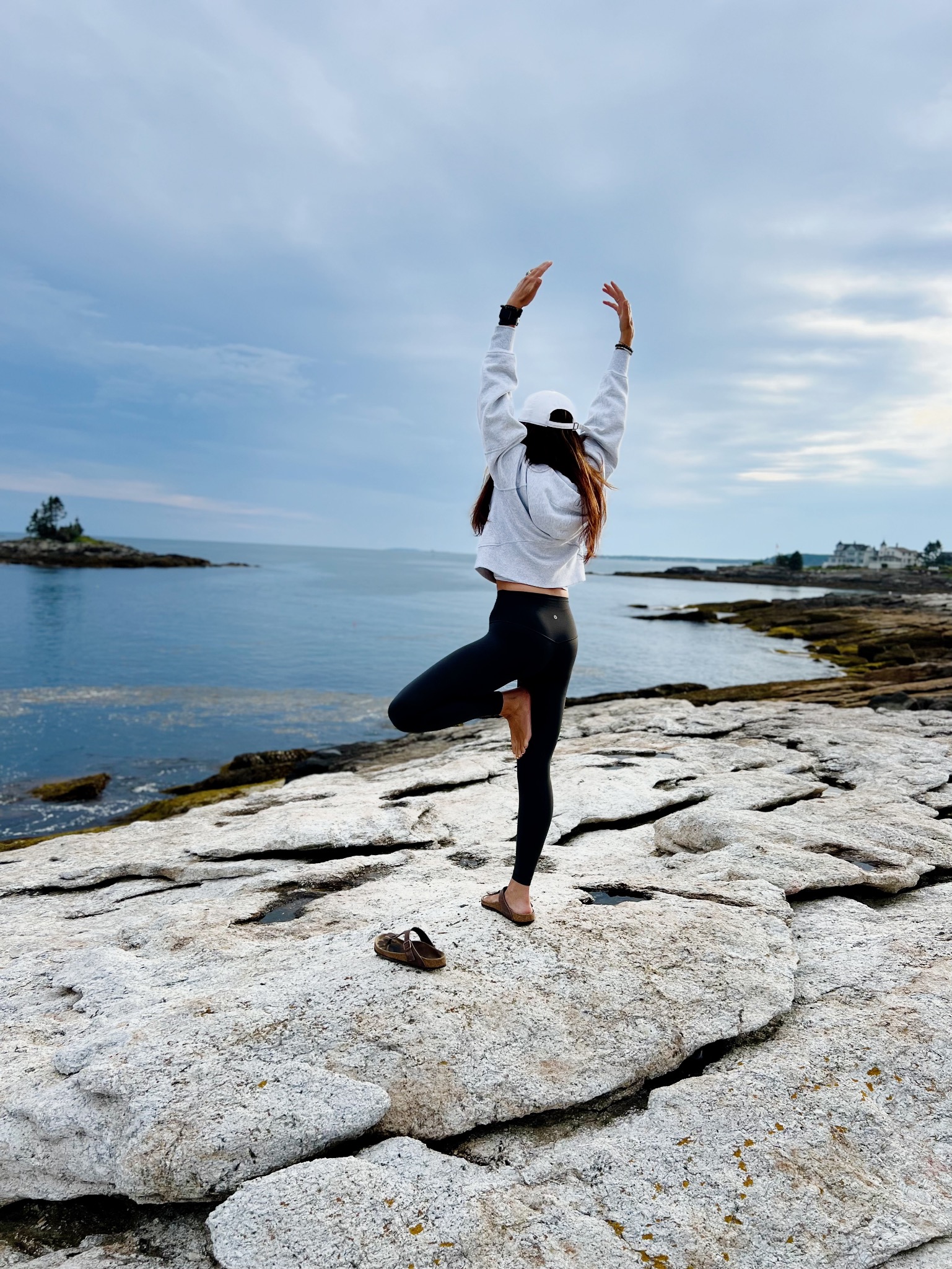 Rebekah practicing tree pose with arms lifted on rocky coastal ledges beside the ocean