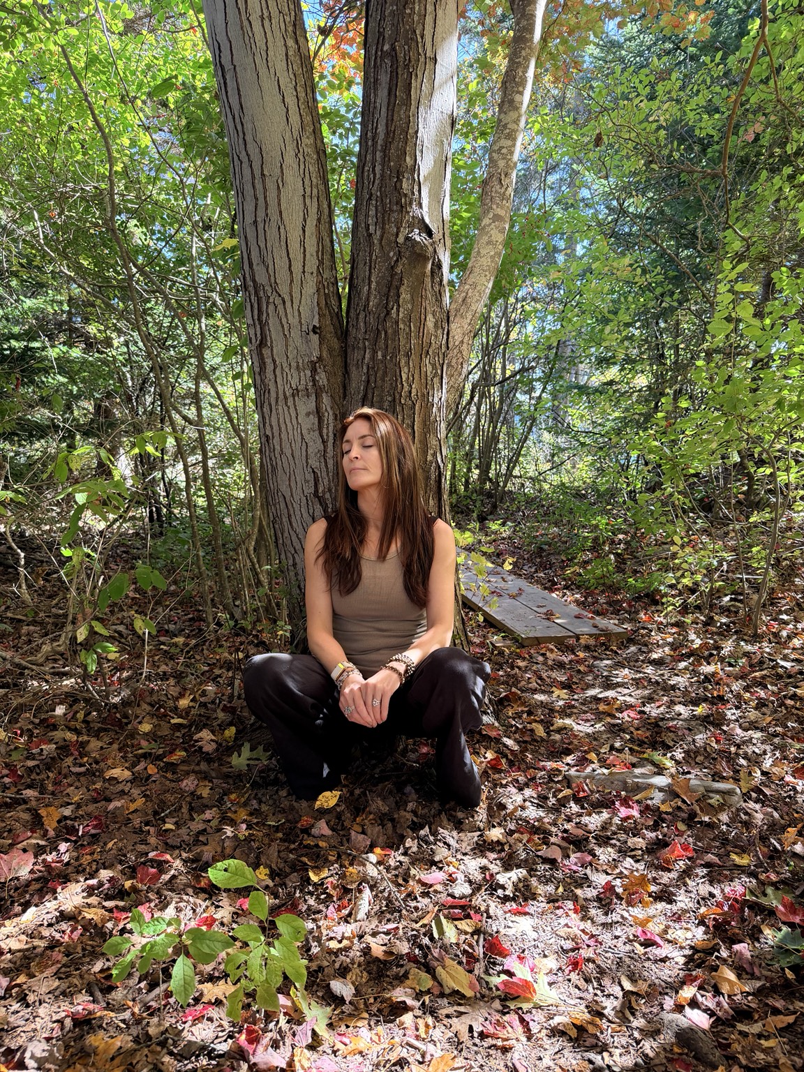 Rebekah seated with eyes closed at the base of a tree, surrounded by autumn leaves and sunlight